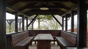 An outdoor pavilion with wooden benches and a central table, surrounded by a wooden structure with a view of trees and a body of water. - Holiday Home Inselperle, Putbus (Putbus)