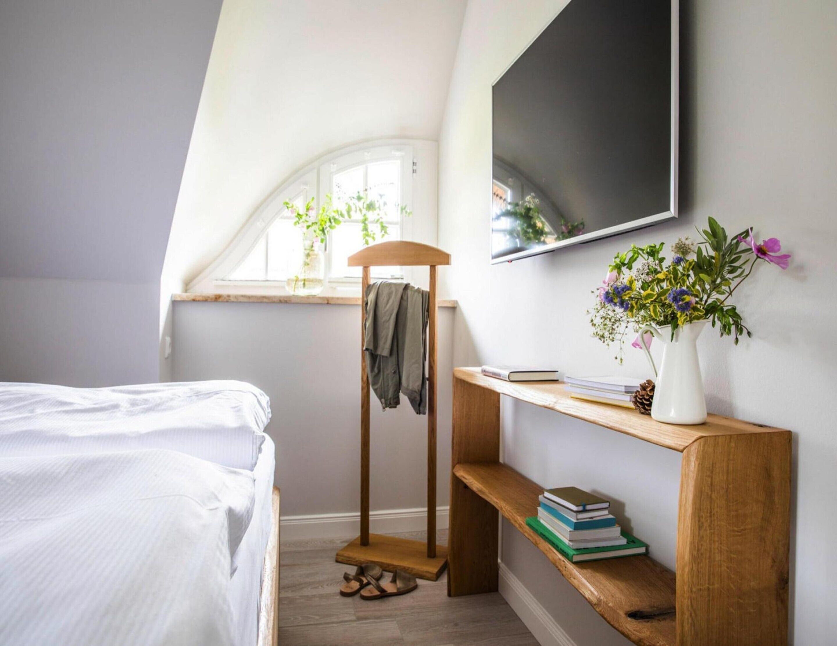 A bedroom with a bed, a wooden shelf with books, a TV on the wall, and a window with plants.