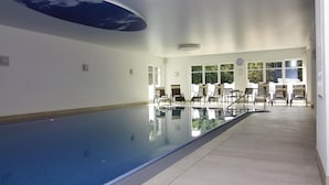 Une piscine intérieure avec des parois en verre transparent, un plafond décoré pour évoquer le ciel et un espace détente avec des chaises et des tables.