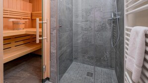 Uma sauna com bancos de madeira e porta de vidro que dá para uma área de chuveiro revestida com azulejos cinzas e um porta-toalhas branco.