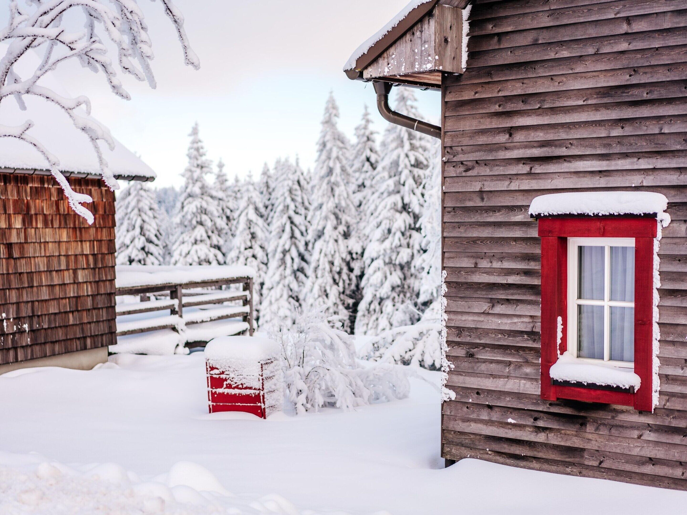 A wooden cabin with a red door and a bench covered in snow.