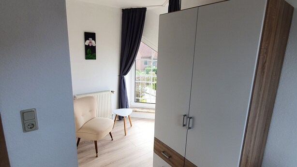 A room with a large wardrobe, a small table, a chair, and a window with curtains. - Ferienhaus in Nessmersiel (Dornum)