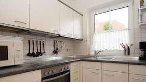 A kitchen with white cabinets, a stove, oven, and a microwave. There is a window with a view of buildings outside. - Ferienhaus in Nessmersiel (Dornum)