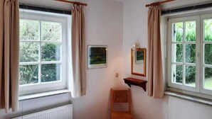 Ein kleiner, ordentlicher Raum mit einem Bett, einem Sessel, einem kleinen Tisch und zwei mit Vorhängen versehenen Fenstern.