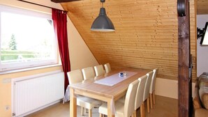 A dining area with a wooden table and chairs, a hanging light fixture, a window with red curtains, and a radiator. - Ferienwohnung Alte Schmiede, Zemitz (Zemitz)