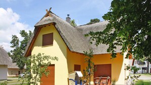 Een traditioneel huis met rieten dak, een klein terras en een tuin met een barbecue en buitenzitplaatsen.
