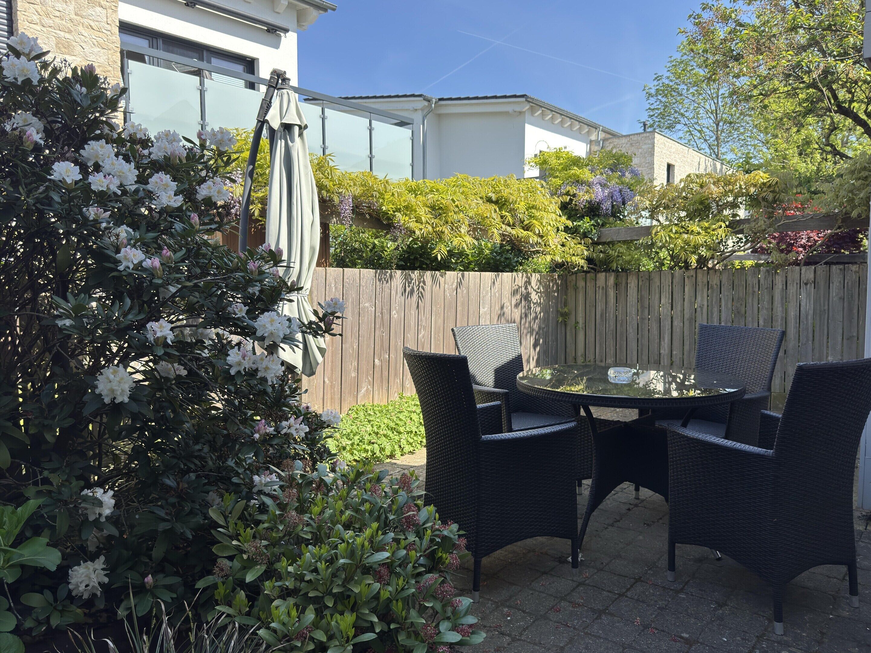 Un patio con mobiliario de mimbre y una mesa de vidrio, rodeado de exuberante follaje y plantas con flores.