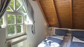 Ein Schlafzimmer mit schrägemausertem Dach, einem fenster mit Vorhängen, einem Bett mit blauer Bettwäsche und einer Nachttischlampe.