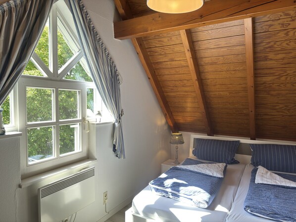 A bedroom with a sloped ceiling, a window with curtains, a bed with blue bedding, and a bedside lamp. - Ferienhaus Fischer Sien Huus, Steinhude (Wunstorf)