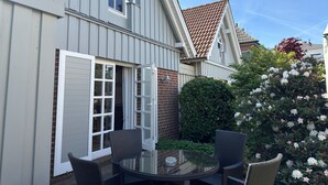 Ein Haus mit einer Terrassenfläche, die einen Glastisch und Korbstühle bietet, umgeben von einer Ziegelmauer und einem Busch mit weißen Blüten.