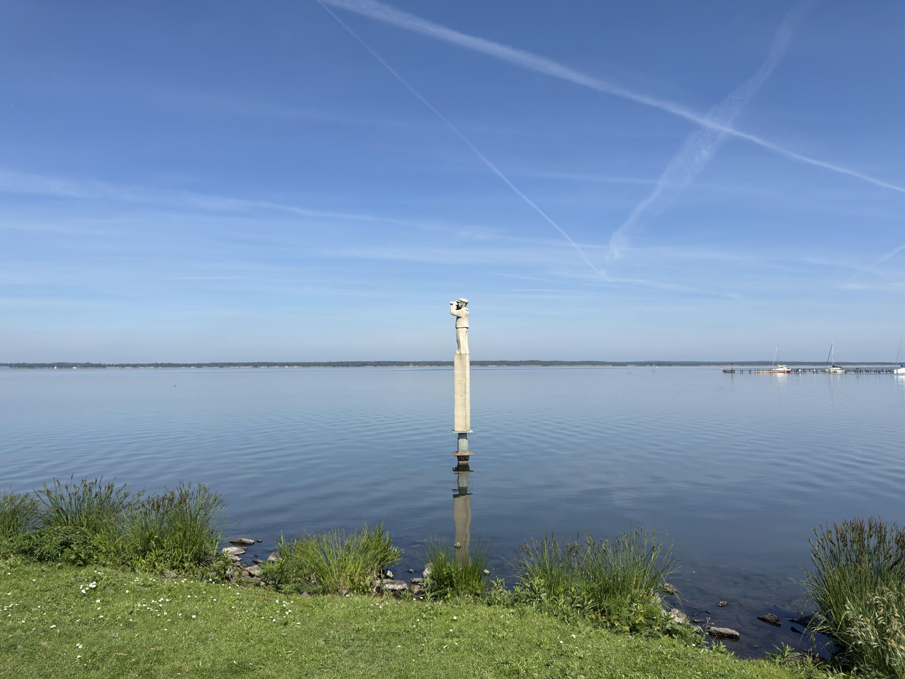 A calm lake with a tall, narrow structure in the center, surrounded by grassy banks and a clear blue sky.