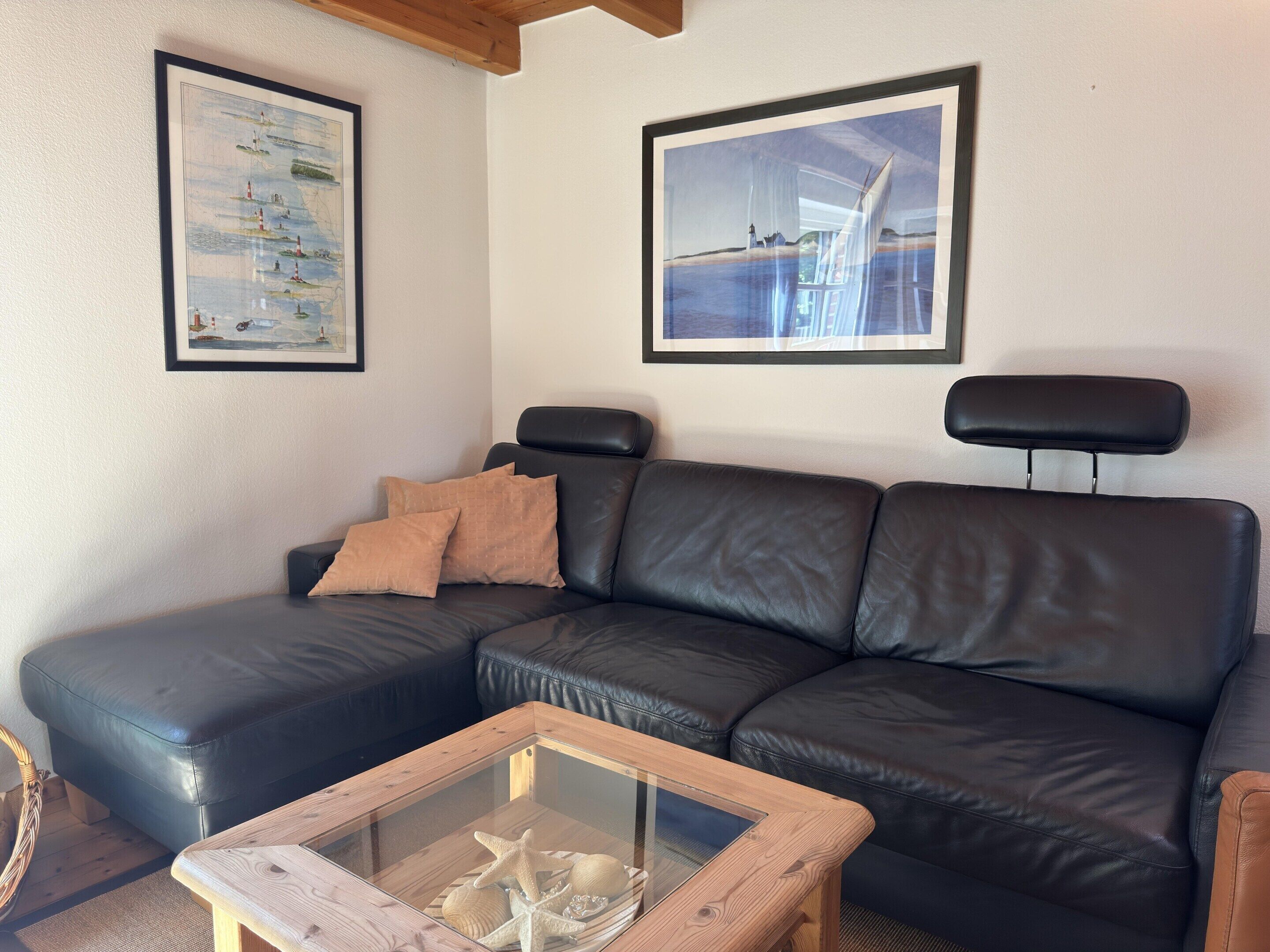 A living room with a black leather sofa, a glass coffee table with decorative items, and two framed artworks on the wall.