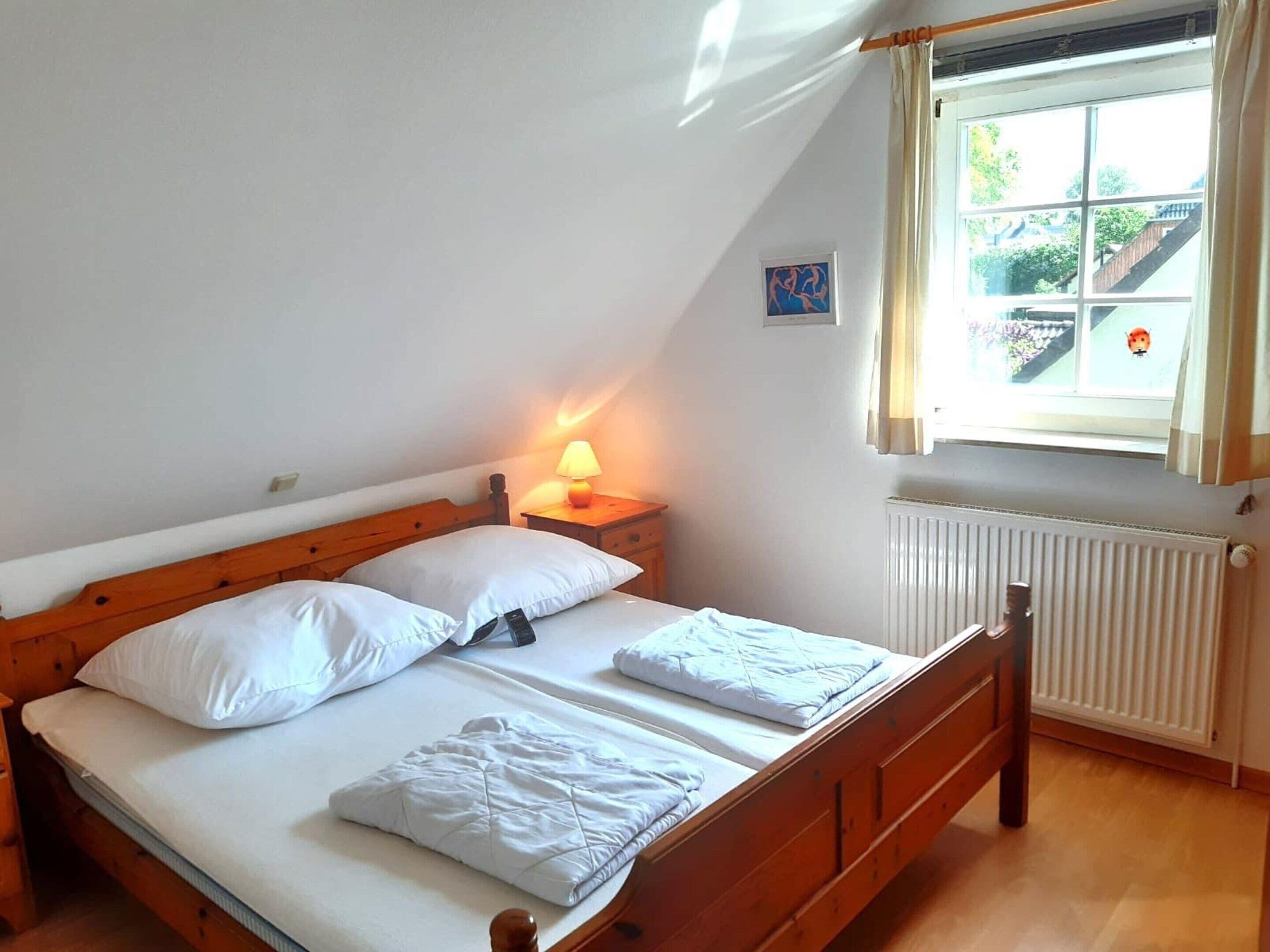 Uma cama de solteiro com lençóis brancos, cabeceira de madeira, mesa de cabeceira com abajur e janela com cortinas.