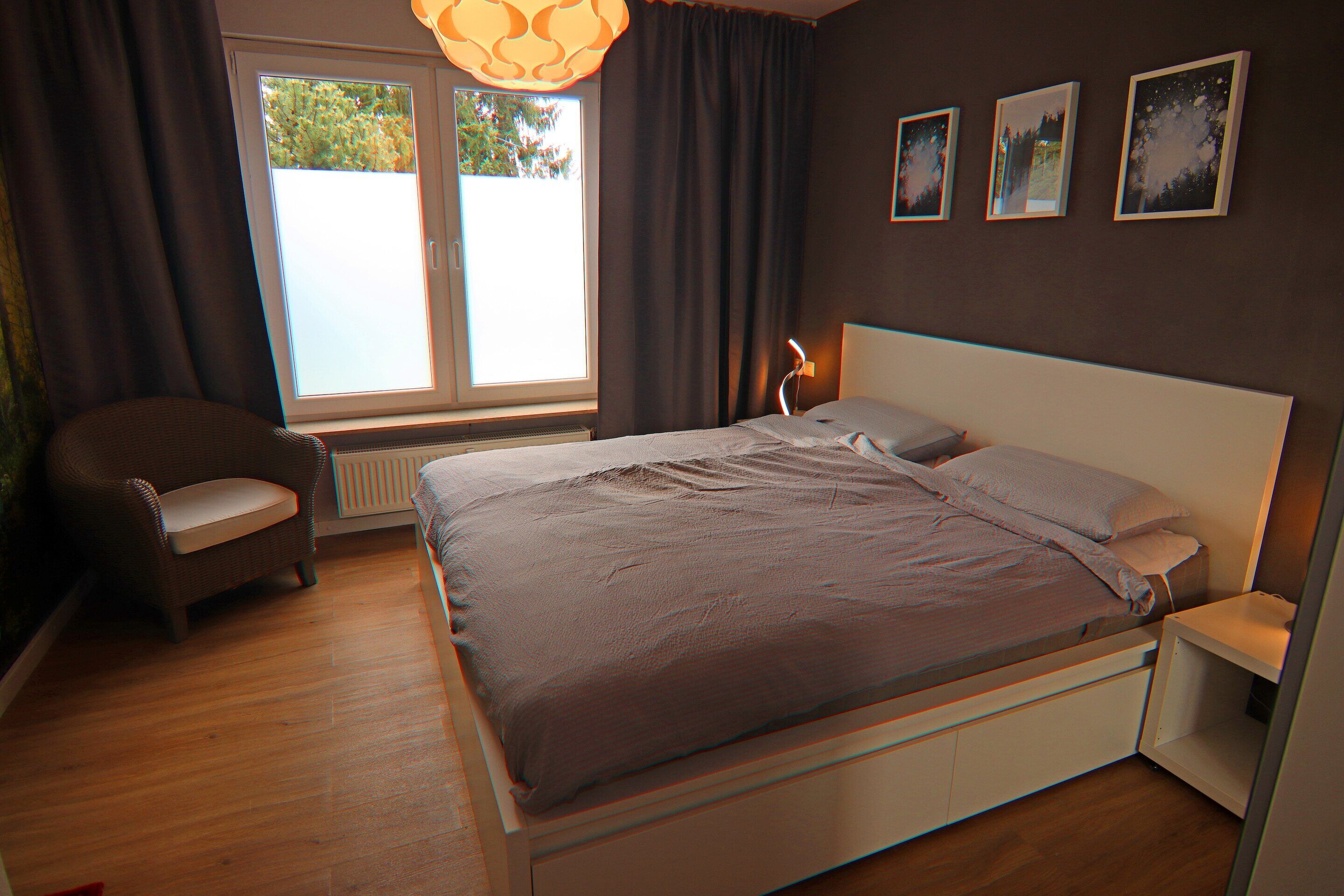 Ein Schlafzimmer mit Bett, Sessel, Nachttisch, Fenster mit Vorhängen und drei gerahmten Bildern an der Wand.