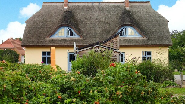 Une maison au toit de chaume et dotée de deux cheminées, entourée d’une végétation luxuriante et de plantes en fleurs.