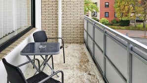 Un balcón con una pequeña mesa y dos sillas, un muro de ladrillo y vistas a un edificio con fachada roja y árboles.