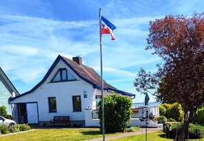 Una casa blanca con techo azul, un asta con bandera, un banco y un pequeño bote.