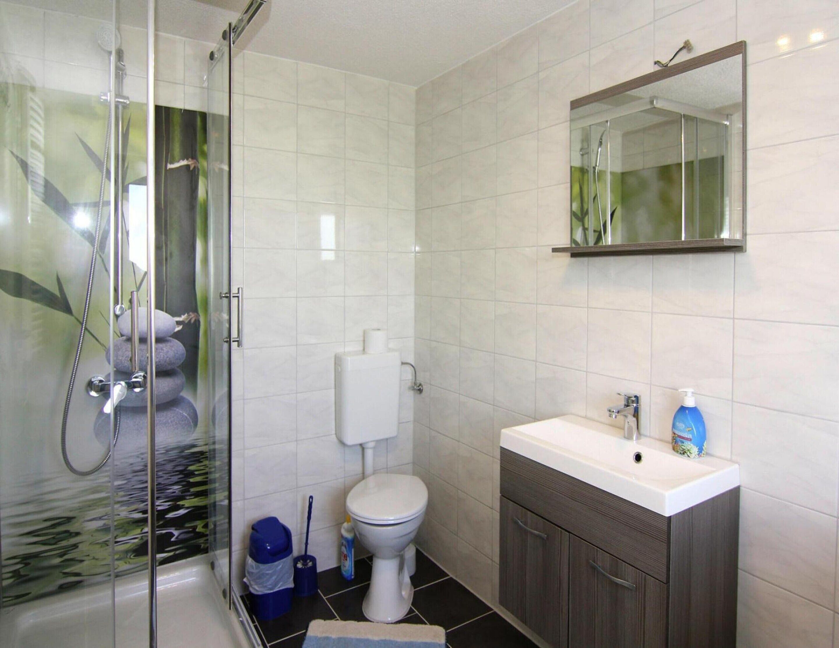 A bathroom with a glass shower enclosure, a toilet, a sink with a mirror, and a blue bottle of hand soap.
