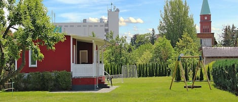 A red building with a white porch, a grassy yard, and a church with a green spire in the background.