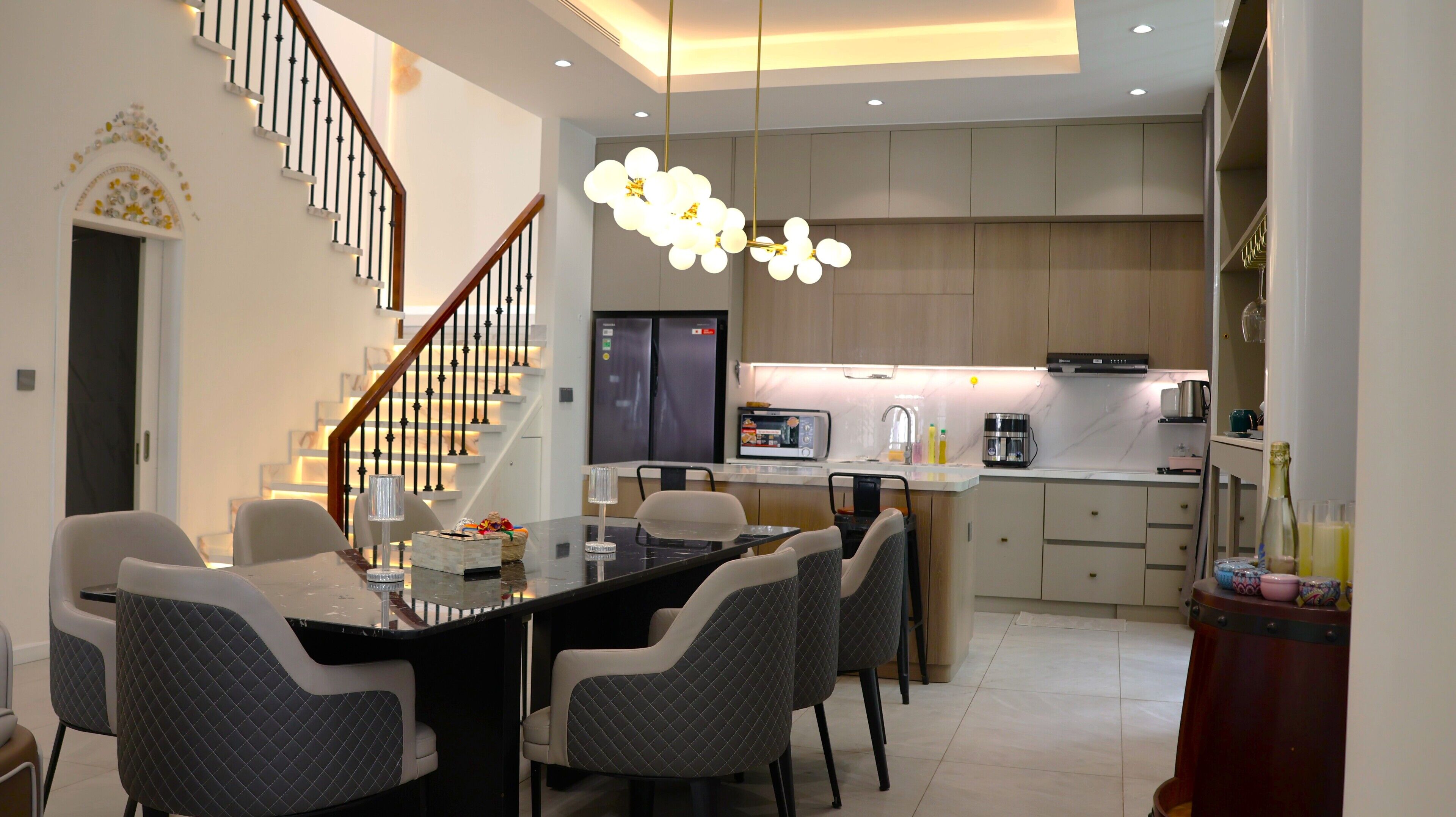 A modern kitchen with a central island, a dining area with a glass table, and a staircase with a railing.