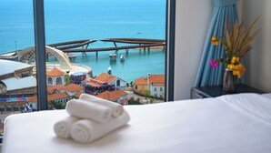 A hotel room with a bed, a window with a view of a coastal city, and a vase with flowers on a nightstand. -  La isla Apartments & Villas – Sea View & Fireworks Phu Quoc (Phu Quoc)