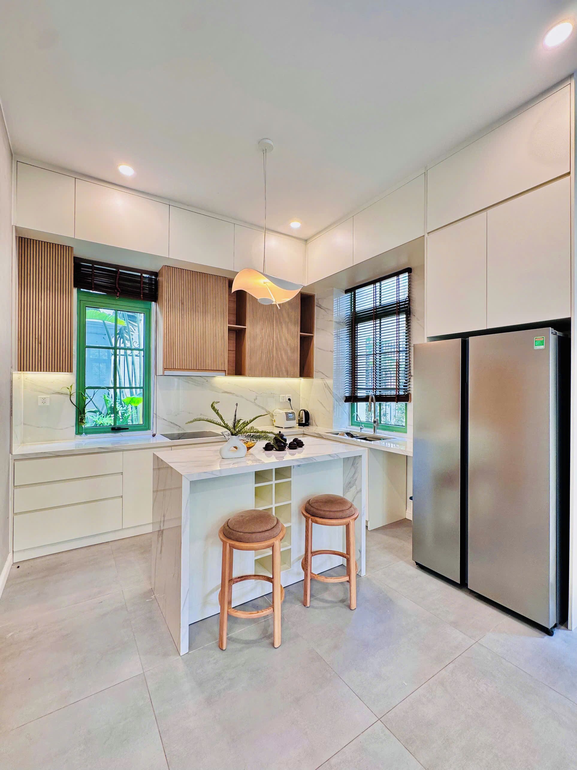 A modern kitchen with a central island, two stools, a refrigerator, and a window with blinds.