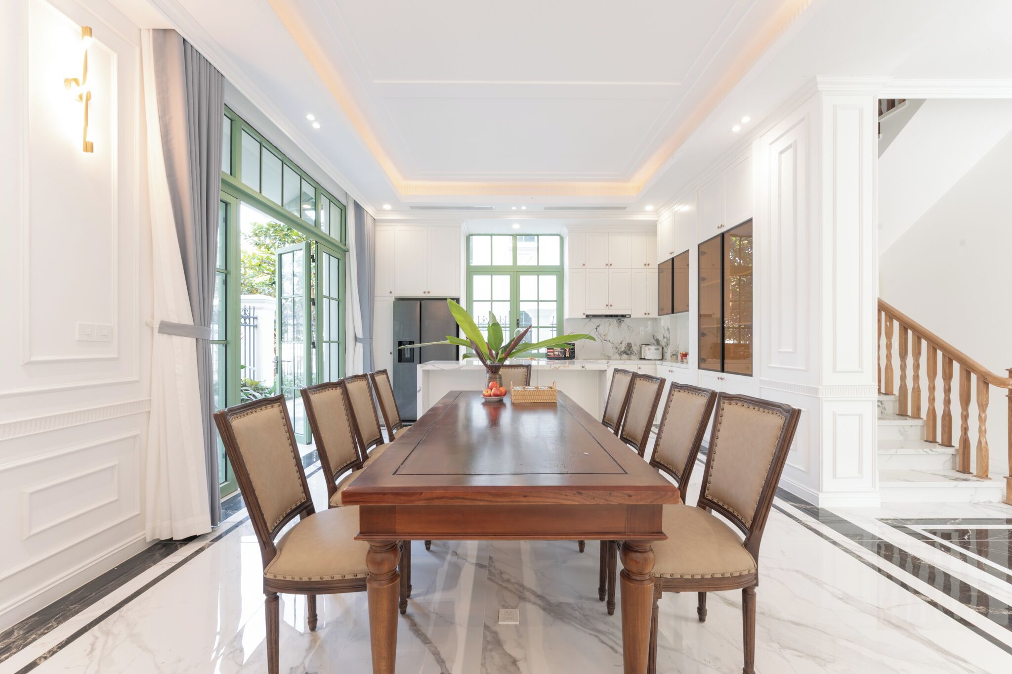 A dining room with a large wooden table, chairs, a vase with plants, and a view of the kitchen and staircase.