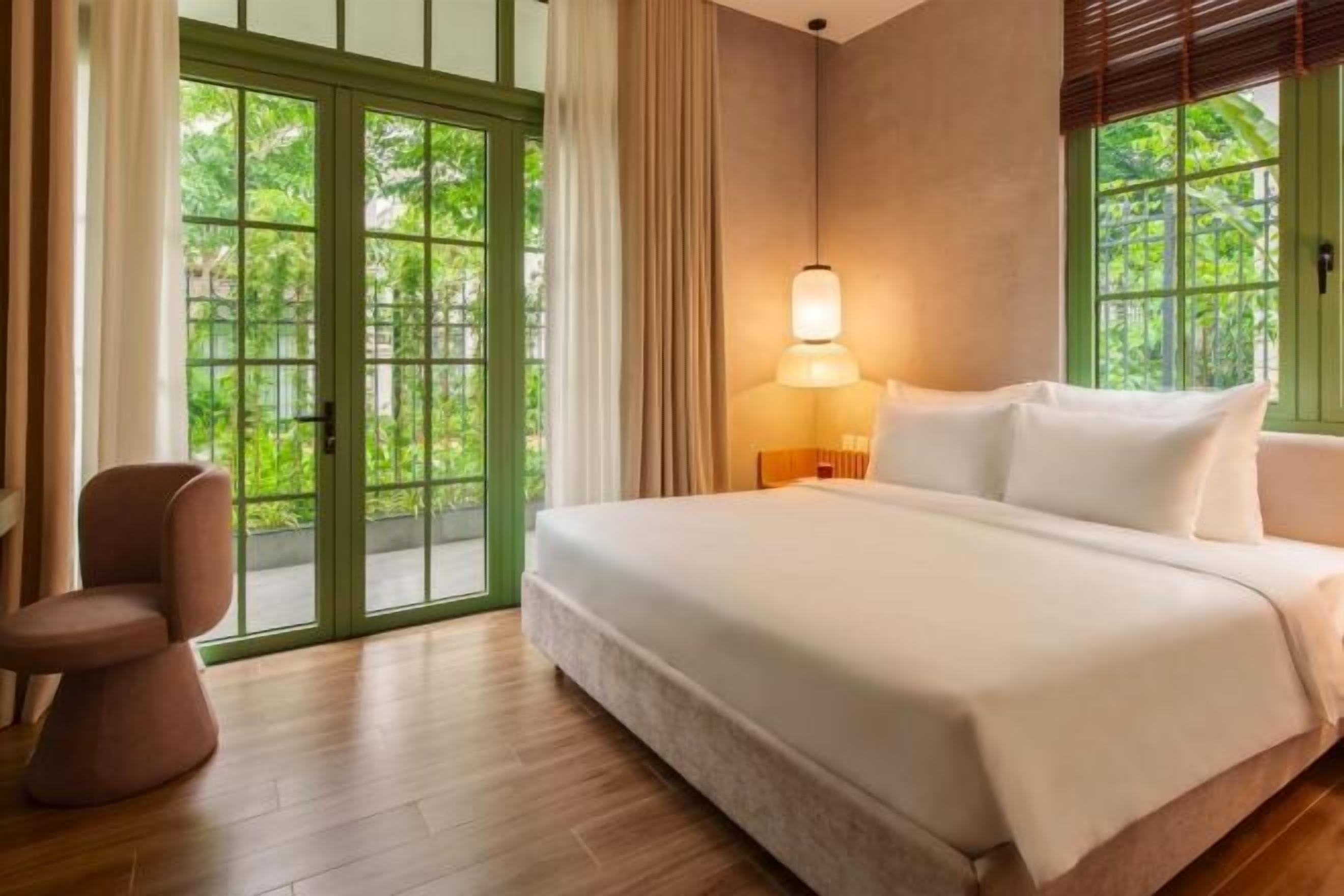 A hotel room with a bed, a chair, a window with curtains, and a door with a view of greenery.