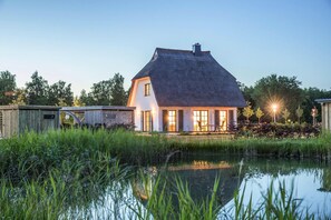 Een huis met rieten dak, een vijver op de voorgrond en een houten tuinhuis aan de linkerkant.
