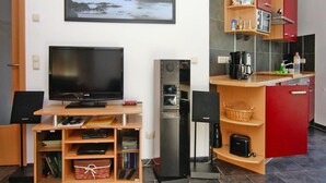 A compact living space with a flat-screen TV, a mini-fridge, a compact kitchen area with a coffee maker, and a bookshelf. - Doppelhaushaelfte am Hafen von Neuendorf (Saal)