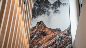 Habitació amb un gran finestral que ofereix vistes a un paisatge de muntanya amb pics rocosos i núvols.