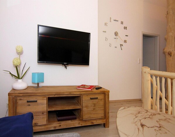 A living room with a wooden TV stand, a flat-screen TV, a vase with flowers, a lamp, and a clock on the wall. - Blockhaus in Trassenheide (Trassenheide)