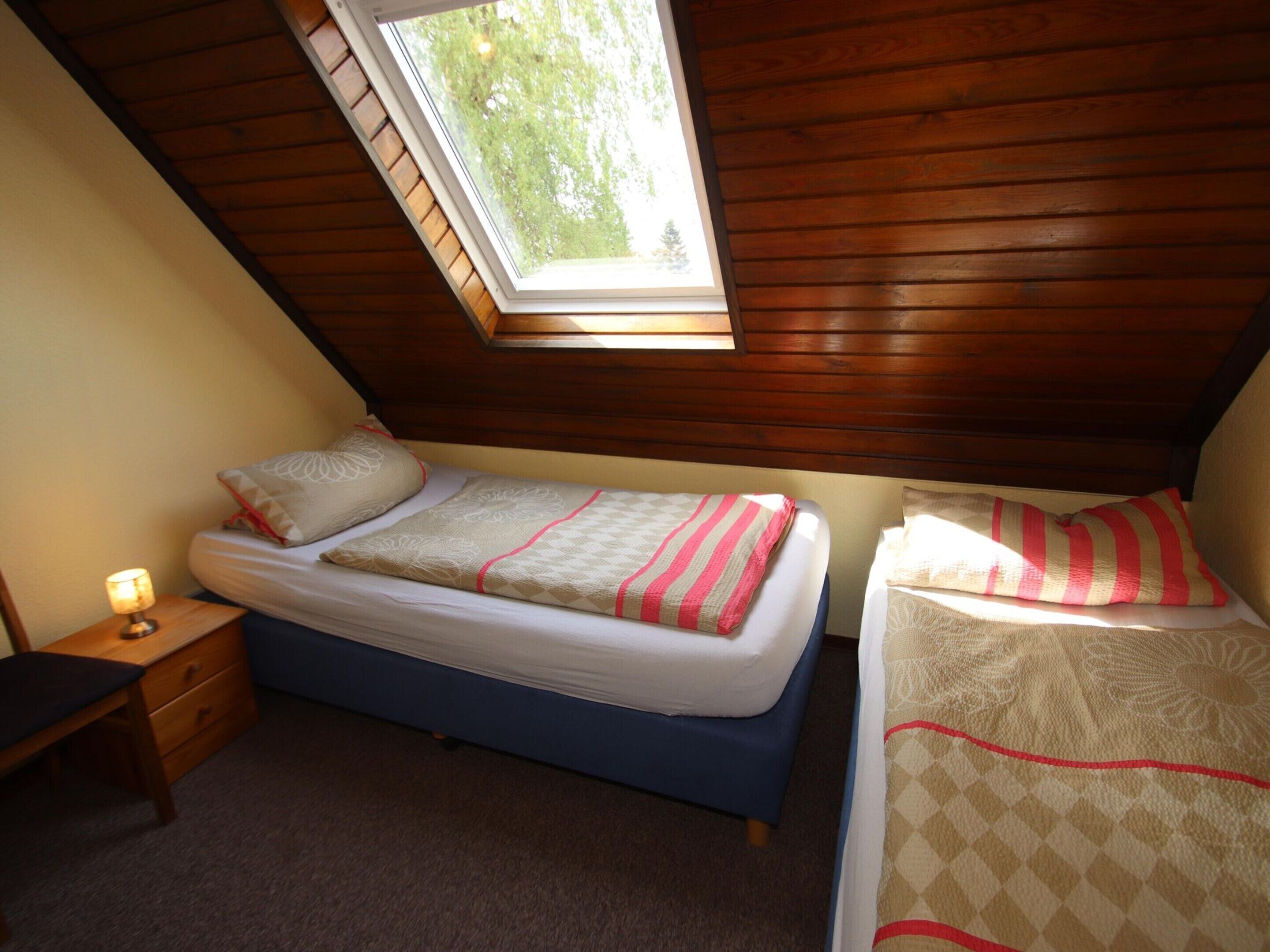 A bedroom with a sloped ceiling and a skylight, featuring two beds with patterned bedding and a bedside table with a lamp.