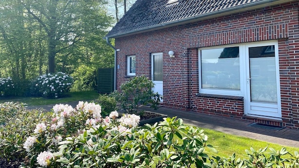Une maison en briques avec un toit en tuiles, deux fenêtres avec volets et un jardin bien entretenu agrémenté d’arbustes en fleurs.
