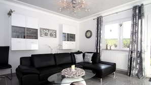A modern living room with a black leather sofa, a glass coffee table, and a chandelier. - Ferienhaus Wiesengrund mit Sauna, Dornumersiel (Dornum)