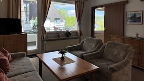Una sala de estar con un sofá, sillones, una mesa de centro y un televisor sobre un mueble bajo. Hay ventanas con cortinas, un radiador y un cuadro enmarcado en la pared.