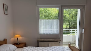Un dormitorio con cama, mesita de noche con lámpara, ventana con vista a follaje y radiador debajo de la ventana.