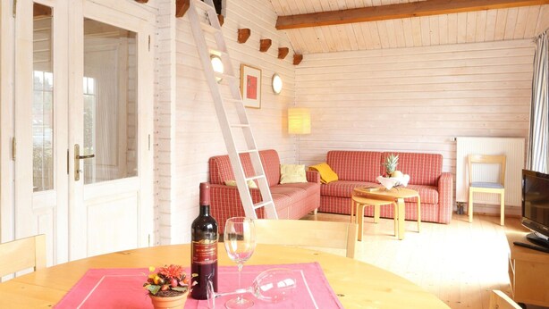 Una habitación acogedora con techo de madera, un sofá rojo, una mesa de madera con una botella de vino y copas, y un televisor.