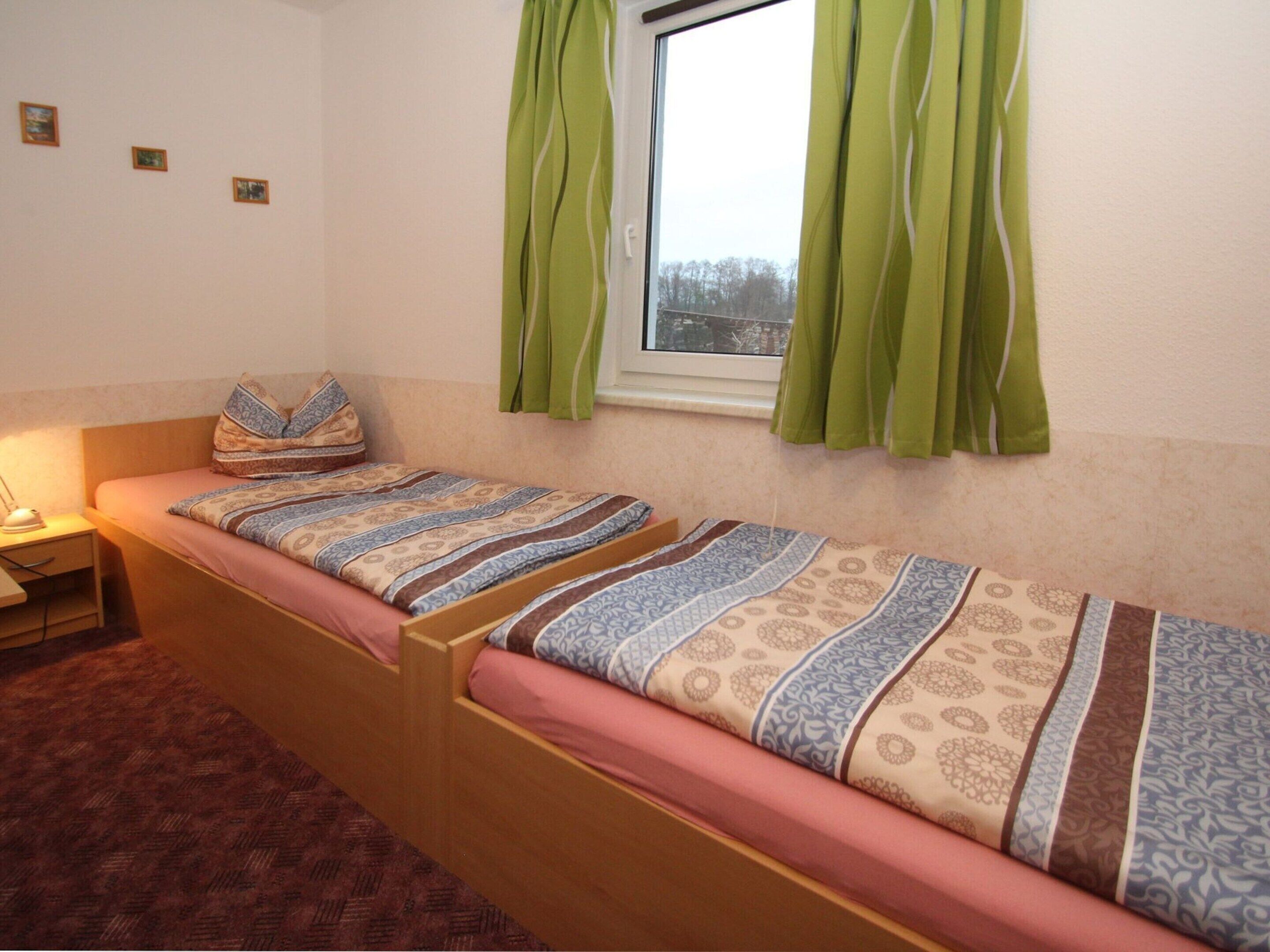 A single-bed room with a wooden frame, a bedside table with a lamp, and a window with green curtains.