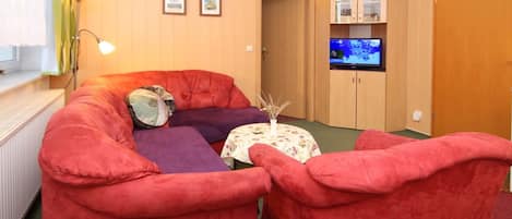 A cozy living room with a red sofa, a purple armchair, and a small round table with a vase. A television is mounted on the wall above a wooden cabinet.