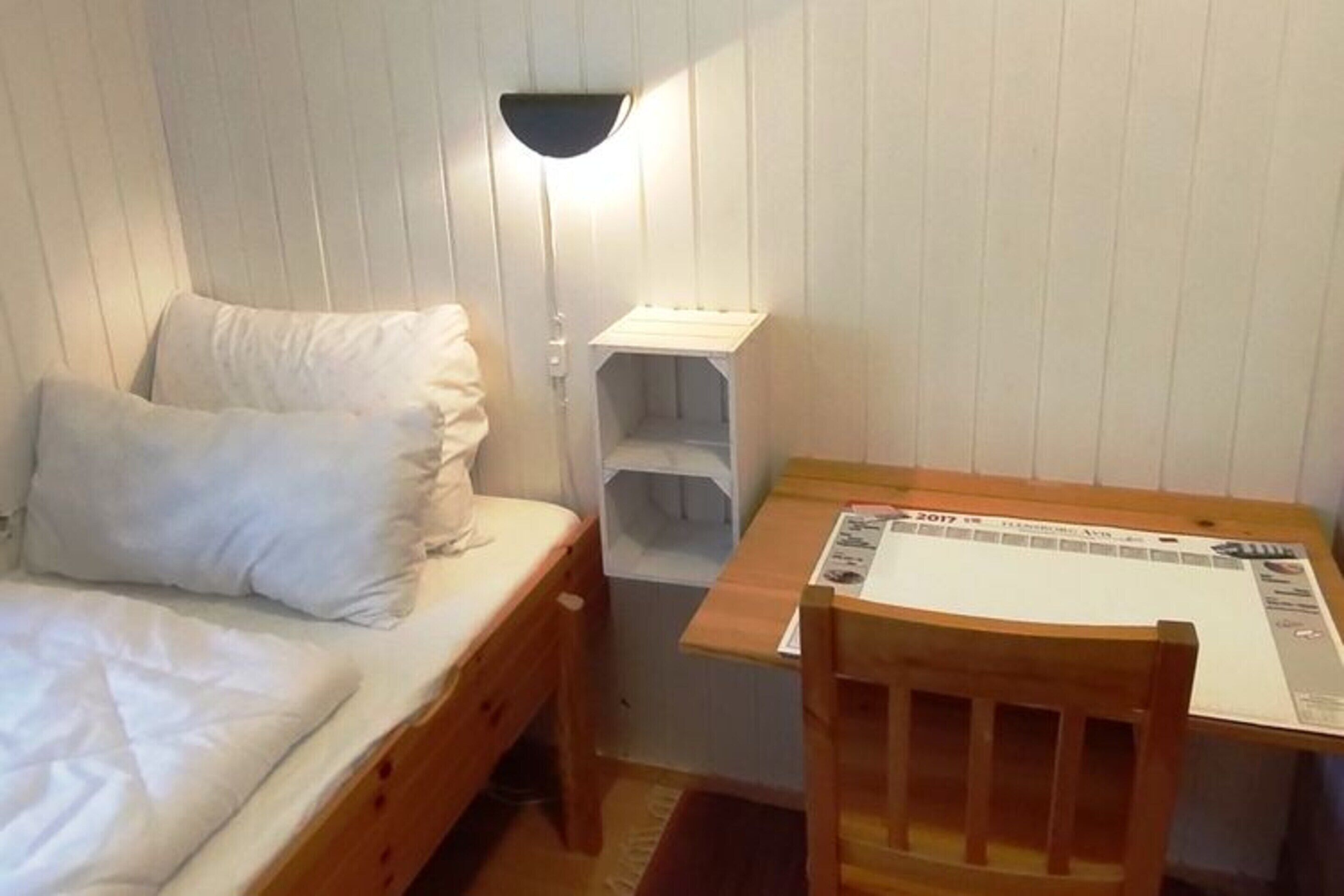 Ein kleiner, gemütlicher Raum mit einem Einzelbett, einem Holz-Nachttisch, einem Schreibtisch mit Stuhl und einer Wandlampe.