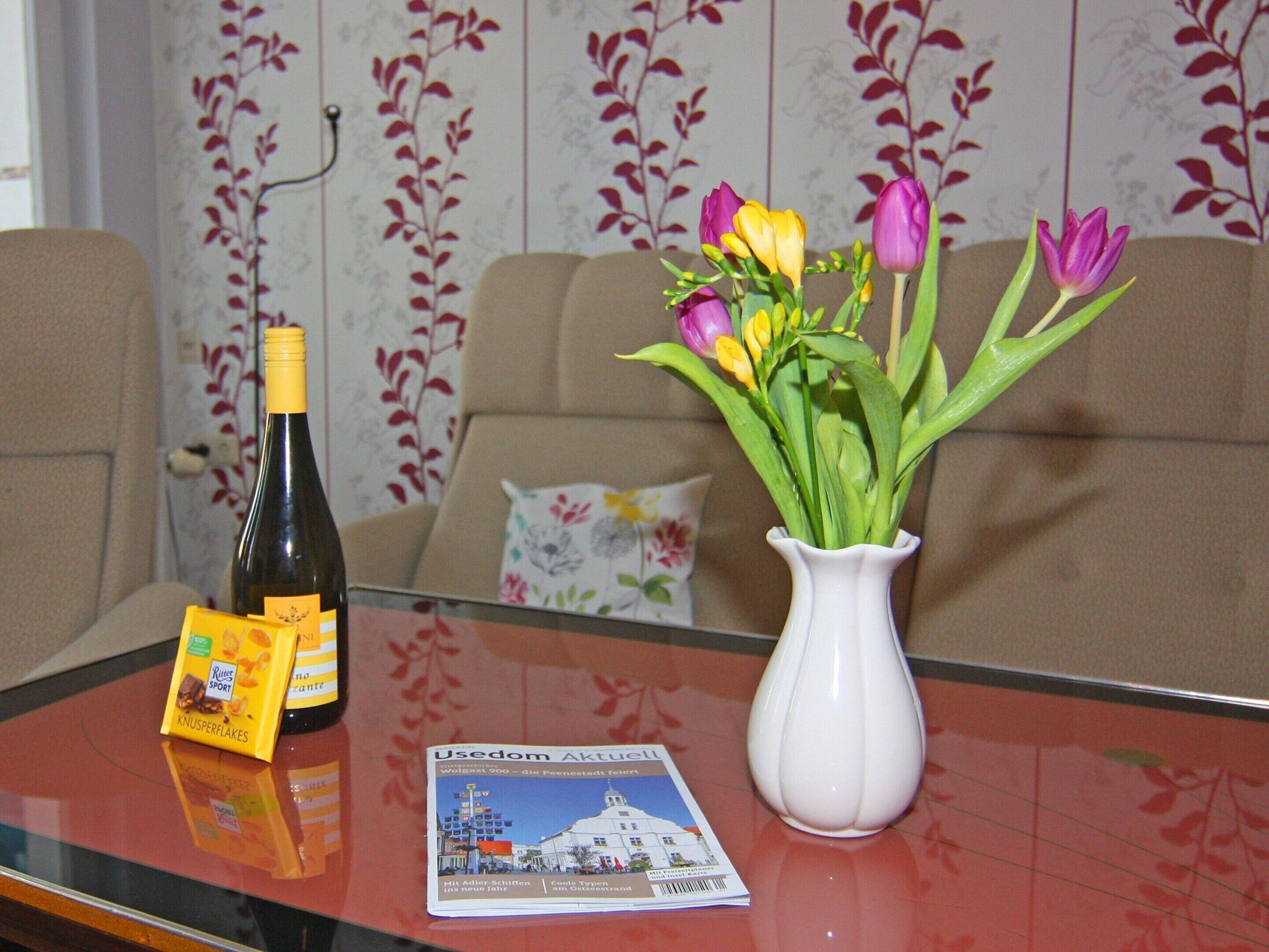 A table with a bottle of wine, a packet of snacks, a brochure, and a vase with flowers.