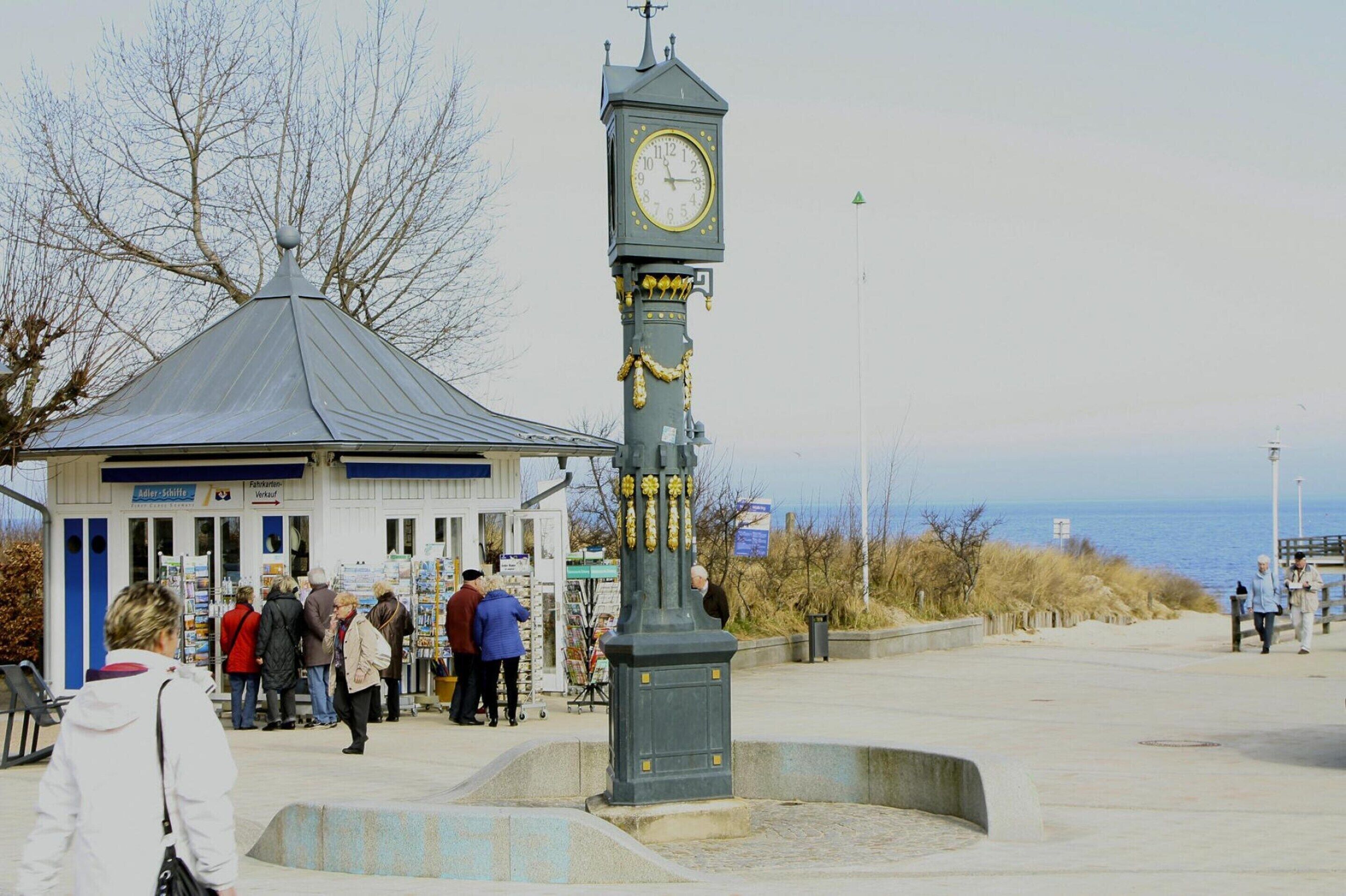 Une tour de l’horloge surmontée d’une girouette, entourée d’une zone pavée avec des bancs et un bâtiment en arrière-plan.