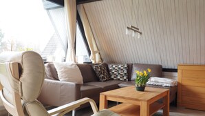 Un salon moderne avec un canapé, un fauteuil, une table basse en bois et une vue sur l’extérieur grâce à de grandes fenêtres.