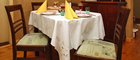 A dining table set for two with a white tablecloth, two chairs, and a small cabinet in the background.