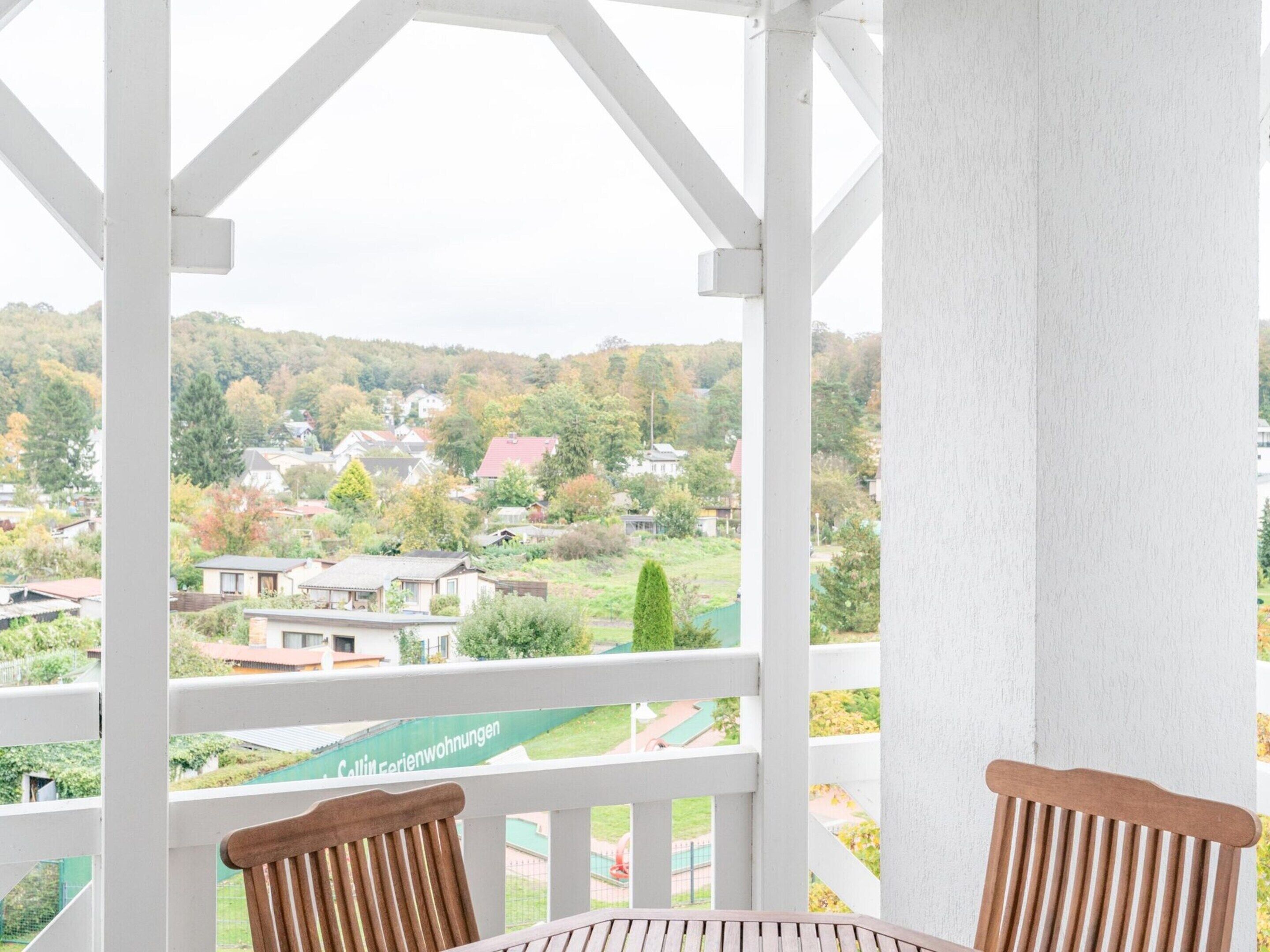 A balcony with a white railing, two wooden chairs, and a view of a residential area with houses and trees.