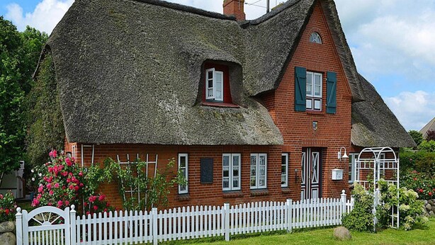 A traditional thatched-roof house with a white picket fence, brick walls, and a small garden with flowering plants. - Ferienwohnung in Oevenum auf Foehr (Oevenum)