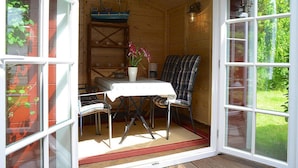 A cozy room with a table and chairs, a small shelf with decorative items, and a view of greenery outside. - Ferienwohnung in Oevenum auf Foehr (Oevenum)