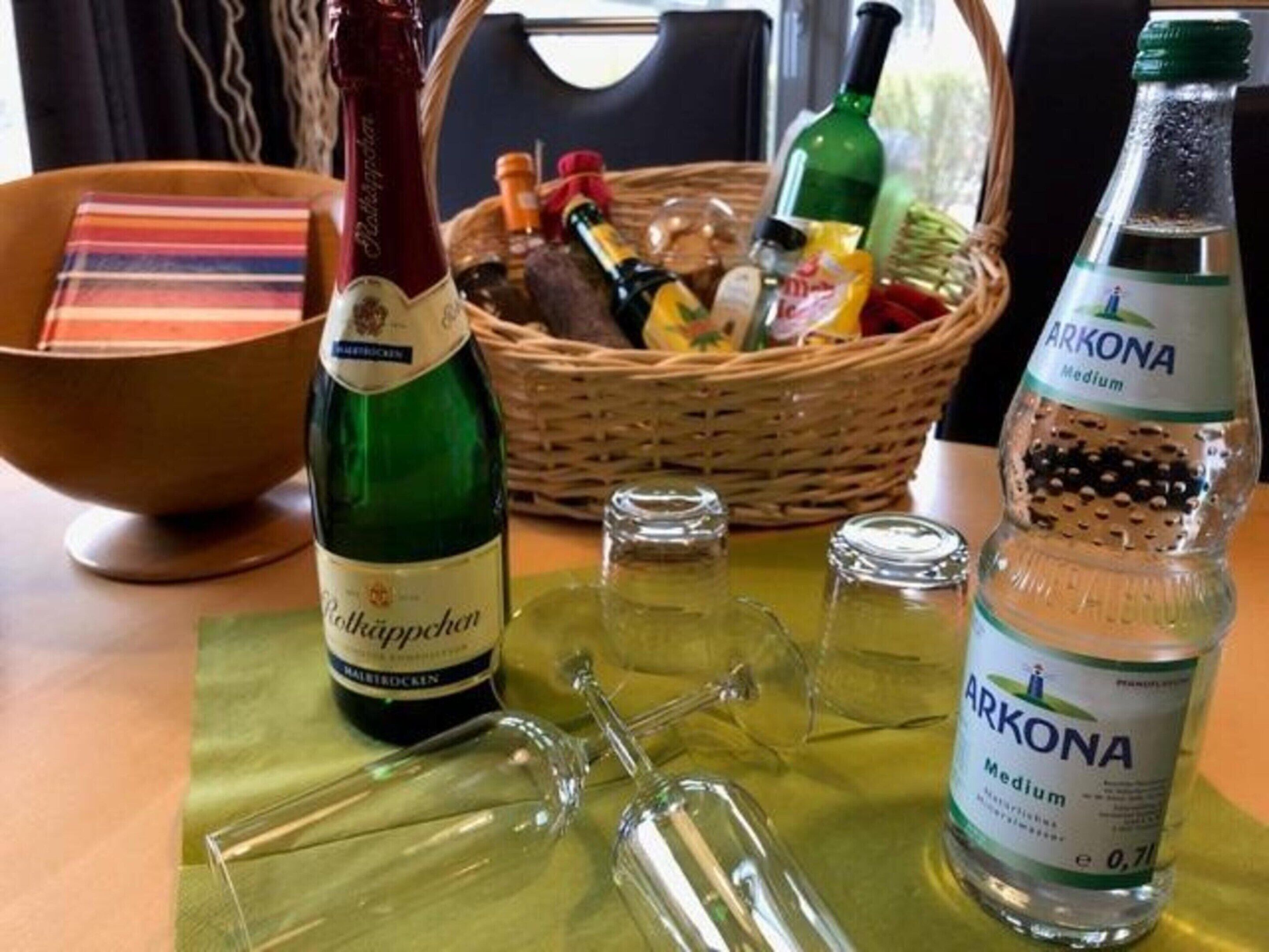 A table with a bottle of Rotkäppchen, a bottle of Arkona medium, and a basket of snacks.