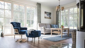 A spacious living room with a large window, a blue armchair, a sofa, a glass coffee table, and a fireplace. - Ferienhaus, Fuhlendorf (Fuhlendorf)